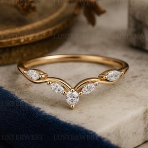 May include: A gold V-shaped ring featuring five marquise-cut white stones set along the band. The band has a polished finish and a curved design that comes to a point at the centre, with the stones arranged symmetrically along the metal frame.
