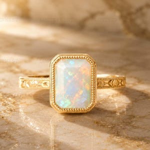 May include: A gold ring featuring a rectangular, emerald-cut opal gemstone with iridescent flashes of color. The stone is set in a beaded gold bezel, and the band displays a textured, engraved pattern along its sides, reflecting light.