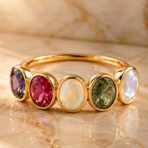 May include: A gold ring featuring five oval-cut gemstones arranged in a row. The stones include a dark purple gem, a vibrant pink gem, a milky white opal, a textured olive green gem, and a translucent white moonstone set in a polished gold band.