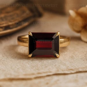 May include: A gold ring featuring a large, rectangular, deep red garnet gemstone set in a four-prong setting. The band is smooth and polished, showcasing a classic design with the dark crimson stone as the central focal point of the jewelry piece.