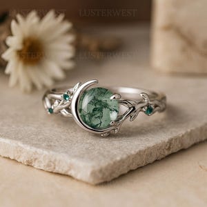 May include: A silver ring featuring a large, round, green moss agate stone set in a crescent moon design. The band has a delicate leaf and vine pattern, with small teal accent stones. The ring is displayed on a neutral surface.