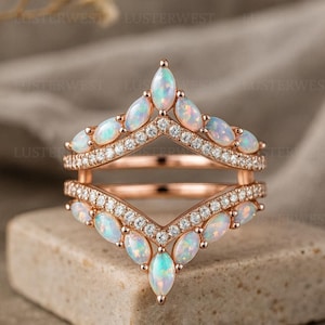Puede incluir: Un protector de anillo en oro rosa con dos bandas en forma de cheurón conectadas en la base. Cada banda está engastada con ópalos de talla marquesa y pequeñas piedras blancas redondas en pavé. El metal tiene un acabado pulido y el diseño crea una abertura en forma de V.
