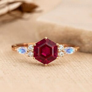 May include: A rose gold ring featuring a large, faceted, hexagonal ruby center stone. Flanking the ruby are smaller white diamonds and pear-shaped moonstones. The ring is set against a neutral background, highlighting the jewelry's sparkle.