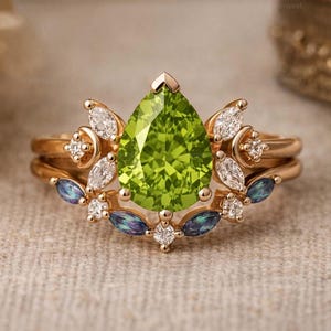 May include: A rose gold ring featuring a large pear-shaped lime green center stone. The setting is adorned with multiple marquise-cut clear white stones and two small marquise-cut iridescent blue-purple stones arranged in a symmetrical floral pattern.