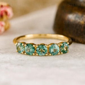 Puede incluir: Un anillo de oro con cinco piedras preciosas redondas de color verde azulado claro. Las piedras están dispuestas en una fila, creando una banda continua de color. La banda del anillo es de oro pulido y las piedras tienen un ligero brillo.