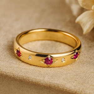 Peut inclure: Une bague en or ornée de trois pierres rouges serties dans des motifs en forme d'étoile, alternant avec de petites pierres claires. La bague est présentée sur une surface neutre, mettant en valeur sa finition polie et ses détails.