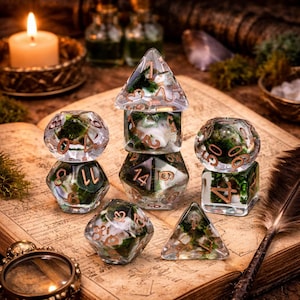 May include: A collection of clear resin dice with green and white inclusions and copper-colored numbering. The dice are arranged on an open book with a candle and other fantasy-themed items in the background. The dice are various shapes.