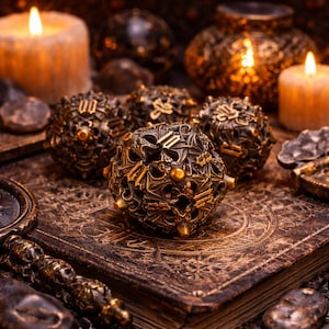 May include: Ornate, bronze-colored spheres with intricate designs rest on a detailed, aged book. Lit candles and other decorative objects create a mystical ambiance. The spheres have a metallic finish and are embellished with small, golden studs.