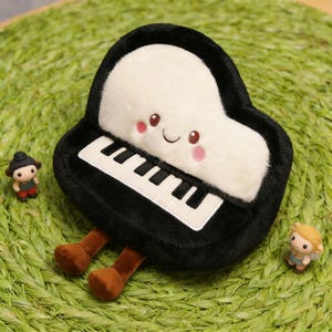 Pode incluir: Um brinquedo de pelúcia de piano com um rosto sorridente, teclas pretas e brancas e pernas marrons. O piano tem um topo branco fofo e é cercado por pequenas figuras em uma superfície verde.