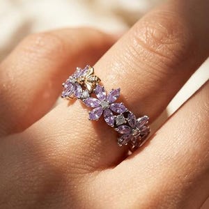 Könnte beinhalten: Ein silberner Ring mit floralem Design. Der Ring hat kleine, lila Edelsteine, die in einem Blumenmuster angeordnet sind. Der Ring wird an einem Finger getragen, mit einem weichen, verschwommenen Hintergrund.