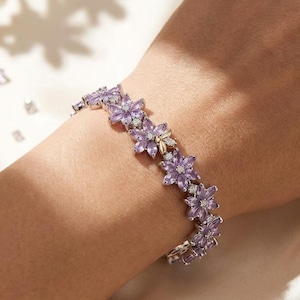 Puede incluir: Un conjunto de pulsera y anillo de plata con un diseño floral. La pulsera y el anillo presentan piedras preciosas moradas en forma de flor y pequeñas piedras blancas. Las joyas se exhiben sobre una superficie blanca.