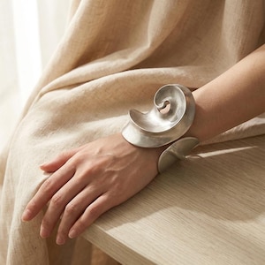 Puede incluir: Un brazalete plateado con un diseño en espiral. El brazalete presenta una gran espiral en capas que se envuelve alrededor de la muñeca, y una pieza curva más pequeña en el otro lado. La joya es una pieza llamativa.
