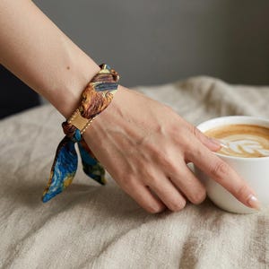 May include: A colorful silk scarf bracelet with a gold-colored clasp and small gold beads. The scarf features a design with shades of blue, brown, and yellow. The bracelet is worn on a wrist.