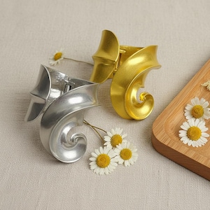 May include: Two cuff bracelets, one silver and one gold, with a swirling, organic design. The bracelets are displayed on a neutral surface, accented with small white and yellow daisies. The gold bracelet is open, revealing the clasp.