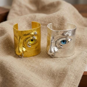 May include: Two cuff bracelets, one gold and one silver, each featuring a sculpted face design. The gold bracelet has a yellow eye, while the silver one has a blue eye. Both have a textured finish and are adorned with small embellishments.