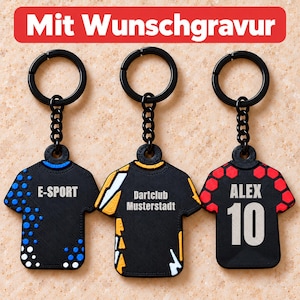 Könnte beinhalten: Drei schwarze Schlüsselanhänger in Trikotform mit schwarzen Ringen. Der linke hat blaue Punkte und die Aufschrift E-SPORT. Der mittlere hat gelbe und weiße Akzente und die Aufschrift Dartclub Musterstadt. Der rechte hat rote Sechsecke, ALEX und 10.