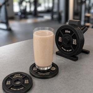 May include: A clear glass filled with a beige protein shake sits on a black coaster shaped like a 25 kg weight plate. Another matching coaster lies nearby, and a small black weight plate rack holds several plates labeled Alex and No Pain No Gain.