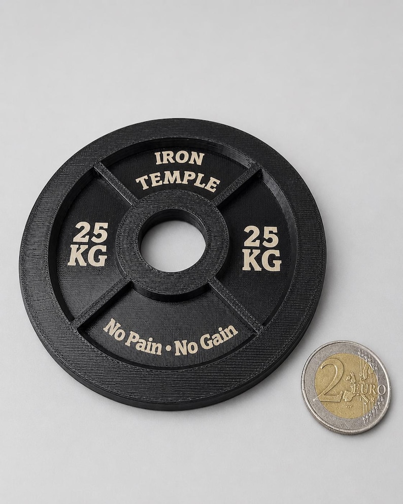 May include: A black circular weight plate with a central hole, featuring the text IRON TEMPLE, 25 KG, and No Pain No Gain in light-colored lettering. A two-euro coin is placed next to the weight plate for scale, showing the relative size.
