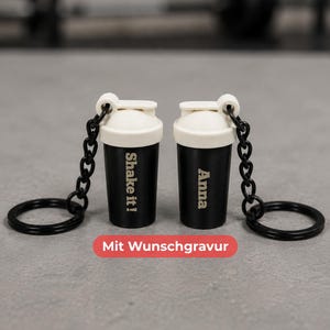 Könnte beinhalten: Zwei schwarz-weiße Mini-Shaker-Schlüsselanhänger mit schwarzen Ketten und Ringen. Ein Shaker trägt den Text Shake it! und der andere den Namen Anna. Ein rotes rechteckiges Etikett unter den Objekten zeigt den Text Mit Wunschgravur.