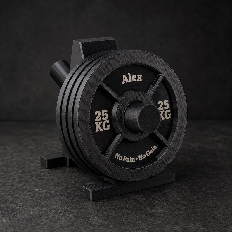 May include: A black weight plate desk accessory featuring the name Alex, the text 25 KG on both sides, and the phrase No Pain &bull; No Gain at the bottom. The circular weight is mounted on a small stand, mimicking the appearance of a gym barbell plate.