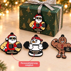 May include: Three flat ornaments featuring a muscular Santa in a red tank top, a snowman lifting weights, and a muscular gingerbread person. The Santa ornament says "Frohe Weihnachten!", the snowman says "Lightweight, buddy!", and the gingerbread person says "Für Max".