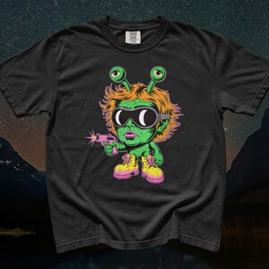 May include: A black t-shirt featuring a graphic illustration of a green alien character with orange hair, wearing black goggles and yellow boots. The alien is holding a pink toy gun that emits a small pink star-shaped flash from the barrel.