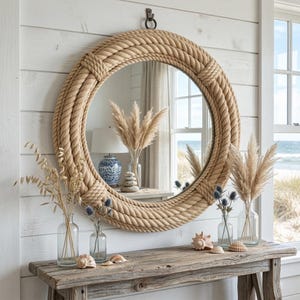 May include: A circular mirror with a rope-style frame is mounted on a white wall. The mirror reflects a coastal view. A wooden console table below displays vases with dried plants, seashells, and blue flowers, creating a seaside theme.