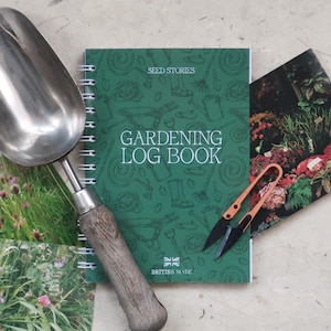 May include: A metal garden scoop with a wooden handle, a green spiral-bound gardening log book with the text "SEED STORIES GARDENING LOG BOOK BRITISH MADE", and a pair of orange-handled snips resting on a floral-patterned paper surface.