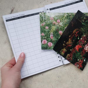 May include: A spiral-bound Bloom & Harvest Tracker planner with a grid layout for tracking plants from January to December. Two rectangular photo inserts are placed on the pages, one showing pink clover flowers and the other a garden display.