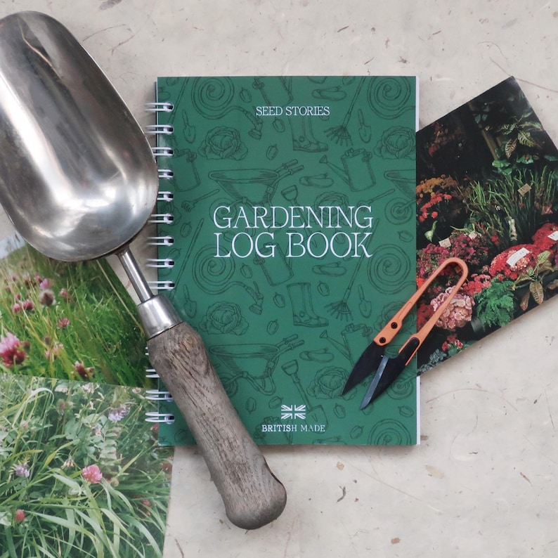 May include: A metal garden scoop with a wooden handle, a green spiral-bound gardening log book with the text "SEED STORIES GARDENING LOG BOOK BRITISH MADE", and a pair of orange-handled snips resting on a floral-patterned paper surface.