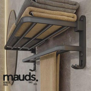 May include: A black metal towel rack with two shelves and hooks, holding folded beige and gray towels. The rack is mounted on a gray tiled wall. The text "mauds. HOMELY FINDS SINCE 2025" is visible.