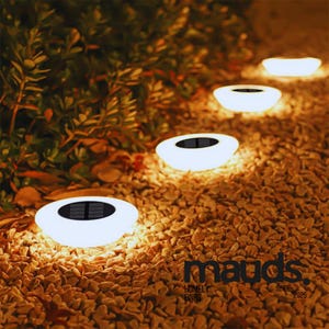 May include: A garden path lit by a series of white, solar-powered lights. Each light features a black solar panel and is set in a bed of small, tan pebbles. Green foliage is in the background. The lights are from "mauds. homely finds since 2025."