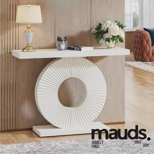 Puede incluir: Consola blanca con diseño circular acanalado. Una estantería blanca sostiene una lámpara, una figura de búho y flores. La mesa tiene el texto "mauds. HOMELY FINDS SINCE 2025".