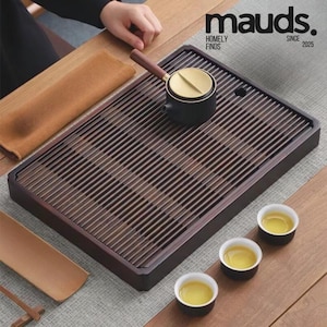 Puede incluir: Una bandeja de té de madera oscura con un diseño de listones, con una pequeña tetera negra con tapa dorada y asa de madera. Tres pequeñas tazas de té llenas de té están dispuestas sobre una superficie de madera. El texto "mauds. HOMELY FINDS SINCE 2025" es visible.