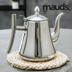 May include: A polished stainless steel teapot with a curved spout and handle, sitting on a woven placemat. The teapot has a lid with a decorative finial. The text "mauds. HOMELY FINDS SINCE 2025" is visible.