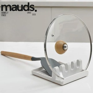 Pode incluir: Um descanso de utensílios de cerâmica branca com uma espátula cinza e uma tampa de vidro transparente com um botão de madeira. A imagem inclui o texto "mauds. HOMELY FINDS SINCE 2025."