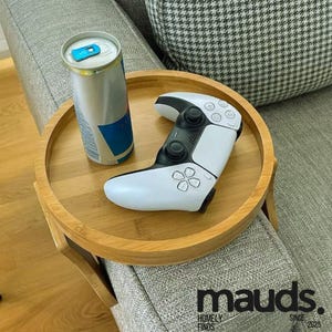 Puede incluir: Una mesa auxiliar redonda de bambú marrón claro con un mando de videojuegos blanco y negro y una lata de bebida energética plateada. La mesa está sobre el brazo de un sofá gris. La mesa tiene el texto "mauds. HOMELY FINDS SINCE 2025".