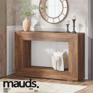 May include: A dark brown wooden console table with a rectangular frame and a hollow centre. A round mirror and two candle holders are on top. A vase with greenery and a basket with pillows and a blanket are also visible. The text "mauds." is at the bottom.