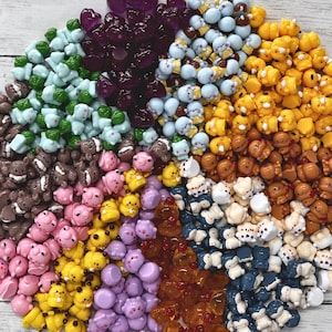 May include: A large collection of small, molded resin cabochons arranged in a circular pattern. The assortment features various colors including purple, light blue, yellow, brown, pink, white, and orange, with diverse shapes and character designs.