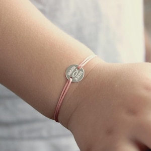 Könnte beinhalten: Ein zartes Armband aus rosa und weißem Kordel mit einem silberfarbenen, kreisförmigen Anhänger. Der Anhänger ist mit dem Namen "Mathilda", einem Fischsymbol und dem Datum "28.02.2018" graviert. Das Armband wird am Handgelenk getragen.