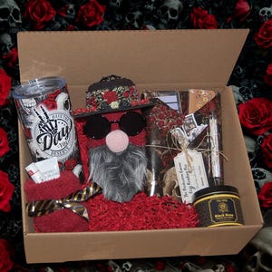May include: A gift box containing a tumbler with a skull and rose print, a gnome figure wearing a hat and round sunglasses, a book wrapped in skull-patterned paper, a black rose scented candle, a lip balm, and a pair of red fuzzy socks.