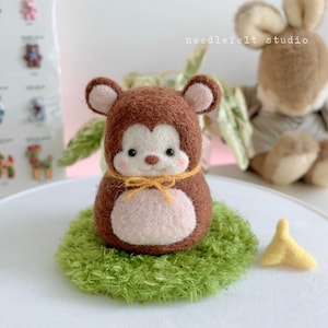 May include: A handcrafted felt monkey figurine with a brown body, light brown face, and large ears. The monkey is sitting on a green, textured base. A yellow bow is around its neck. A small yellow banana is next to the monkey.