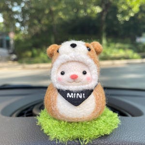 May include: A handcrafted felt figurine of a character wearing a brown bear hat and a black scarf with "MINI" printed on it. The figurine is placed on a green, textured base. The background is blurred, with trees and a road visible.