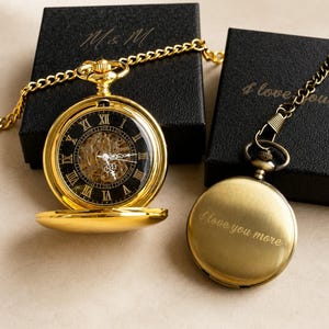Peut inclure: Deux montres de poche dorées avec chaînes. Une montre est ouverte, révélant un cadran noir avec des chiffres romains et des engrenages internes visibles. L'autre montre est fermée, avec le texte gravé I love you more sur sa surface.