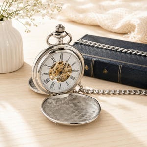 May include: A silver-toned mechanical pocket watch with a white face, black Roman numerals, and an exposed gold-colored gear movement. The watch features a hinged cover with a textured interior and is attached to a matching silver-toned metal chain.