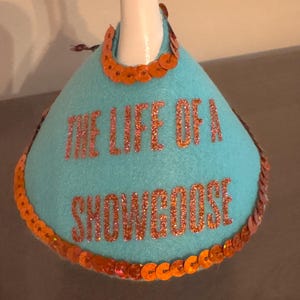 Peut inclure: Un bouchon de bouteille en feutre turquoise avec le texte "THE LIFE OF A SNOWGOOSE" en lettres orange paillet&eacute;es. La garniture est compos&eacute;e de paillettes orange et d'un col en paillettes orange.