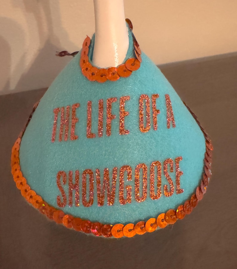 Peut inclure: Un bouchon de bouteille en feutre turquoise avec le texte "THE LIFE OF A SNOWGOOSE" en lettres orange paillet&eacute;es. La garniture est compos&eacute;e de paillettes orange et d'un col en paillettes orange.