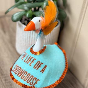 Peut inclure: Figurine d'oie fantaisiste avec un corps blanc, un bec et des pieds orange, et une cape bleue. La cape est orn&eacute;e de paillettes orange et du texte "THE LIFE OF A SHOWGOOSE". L'oie a des plumes orange et bleues.