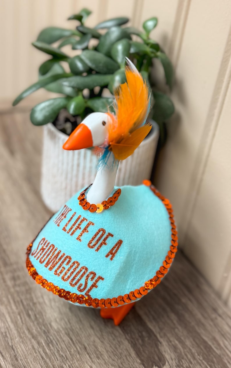 Peut inclure: Figurine d'oie fantaisiste avec un corps blanc, un bec et des pieds orange, et une cape bleue. La cape est orn&eacute;e de paillettes orange et du texte "THE LIFE OF A SHOWGOOSE". L'oie a des plumes orange et bleues.