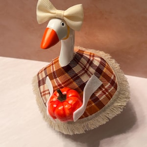 May include: A white goose figurine wearing a cream-coloured ribbon bow on its head and a brown, orange, and white plaid cape with fringe trim. The goose is holding a small, bright orange pumpkin with a dark stem in its white wing-like appendages.